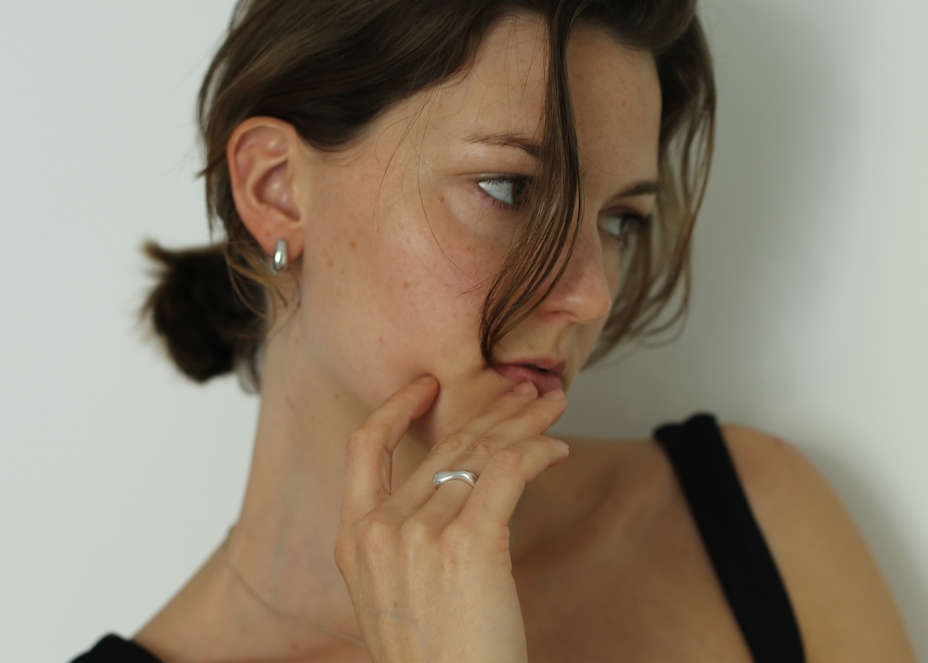 Woman in a black tank top and silver jewelry with hand near mouth against a white background