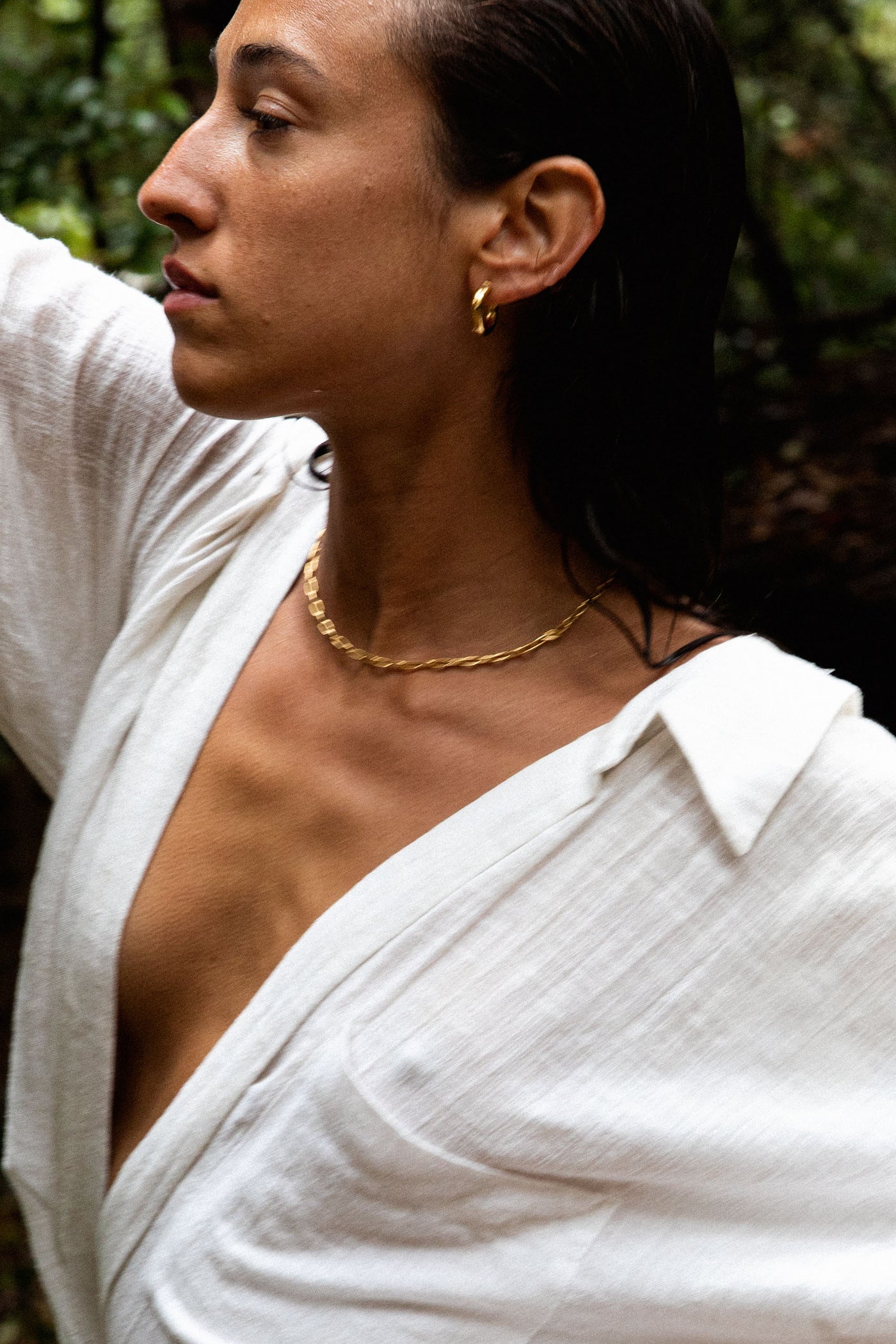 Side profile of a woman wearing a gold chain necklace and small hoop earrings, dressed in a white, open-collar top. She has her arm raised, with a natural outdoor background