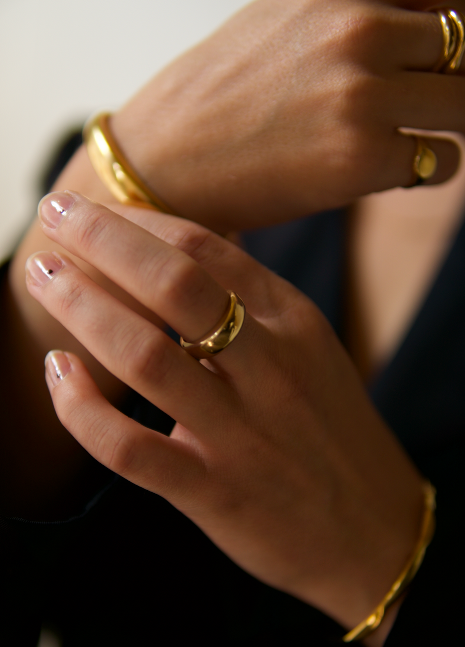 Detail shot of a woman's hands adorned with minimalist gold rings and a matching bracelets, wearing a black blazer.